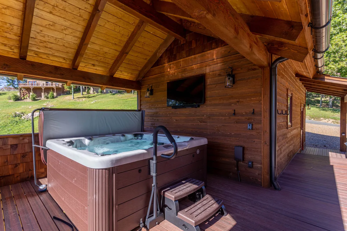 This image captures a stylish outdoor deck featuring a hot tub surrounded by wooden structure, lush greenery, and an outdoor television. The inviting space is perfect for relaxing in the bubbling hot tub or entertaining guests while enjoying movies under the stars.
