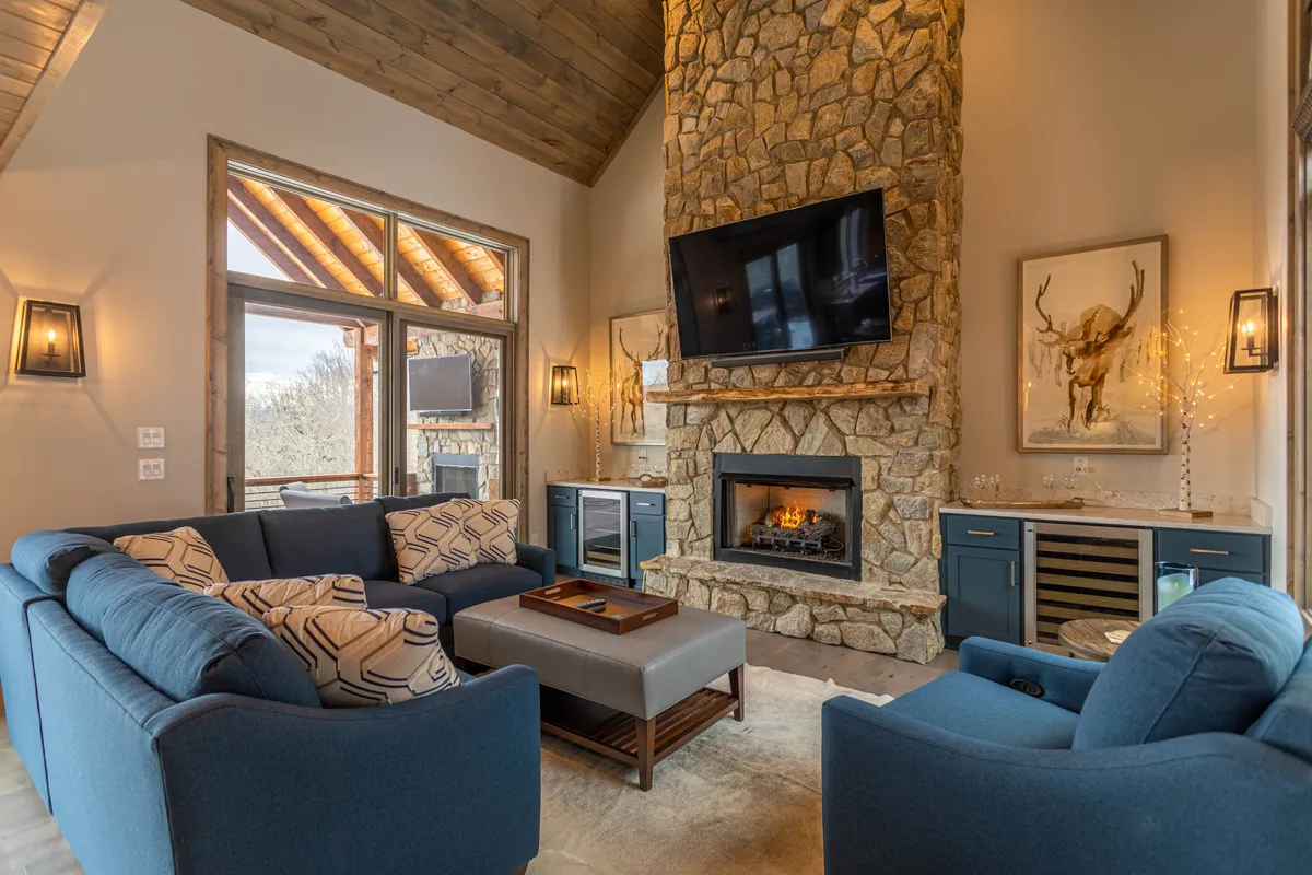 A cozy living room with a stone fireplace, comfortable seating, a large television mounted above the fireplace, and expansive windows offering mountain views. The scene invites relaxation, socialization, and entertainment, with a touch of elegance and warmth.