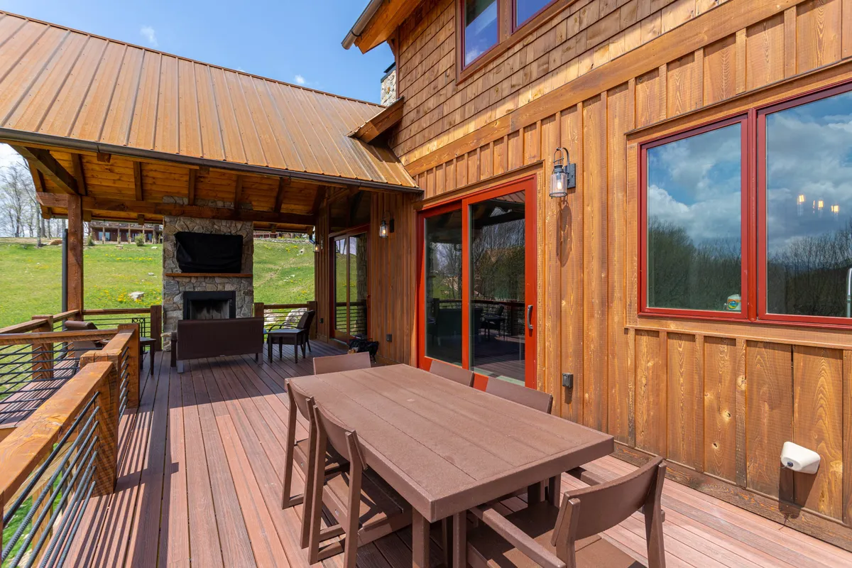 A spacious outdoor patio featuring a dining table set beside a stone fireplace, framed by wooden siding and large windows. The setting is perfect for groups to gather for dinner, enjoy drinks by the fire, or stargaze in comfort, all while remaining immersed in the surrounding natural beauty.