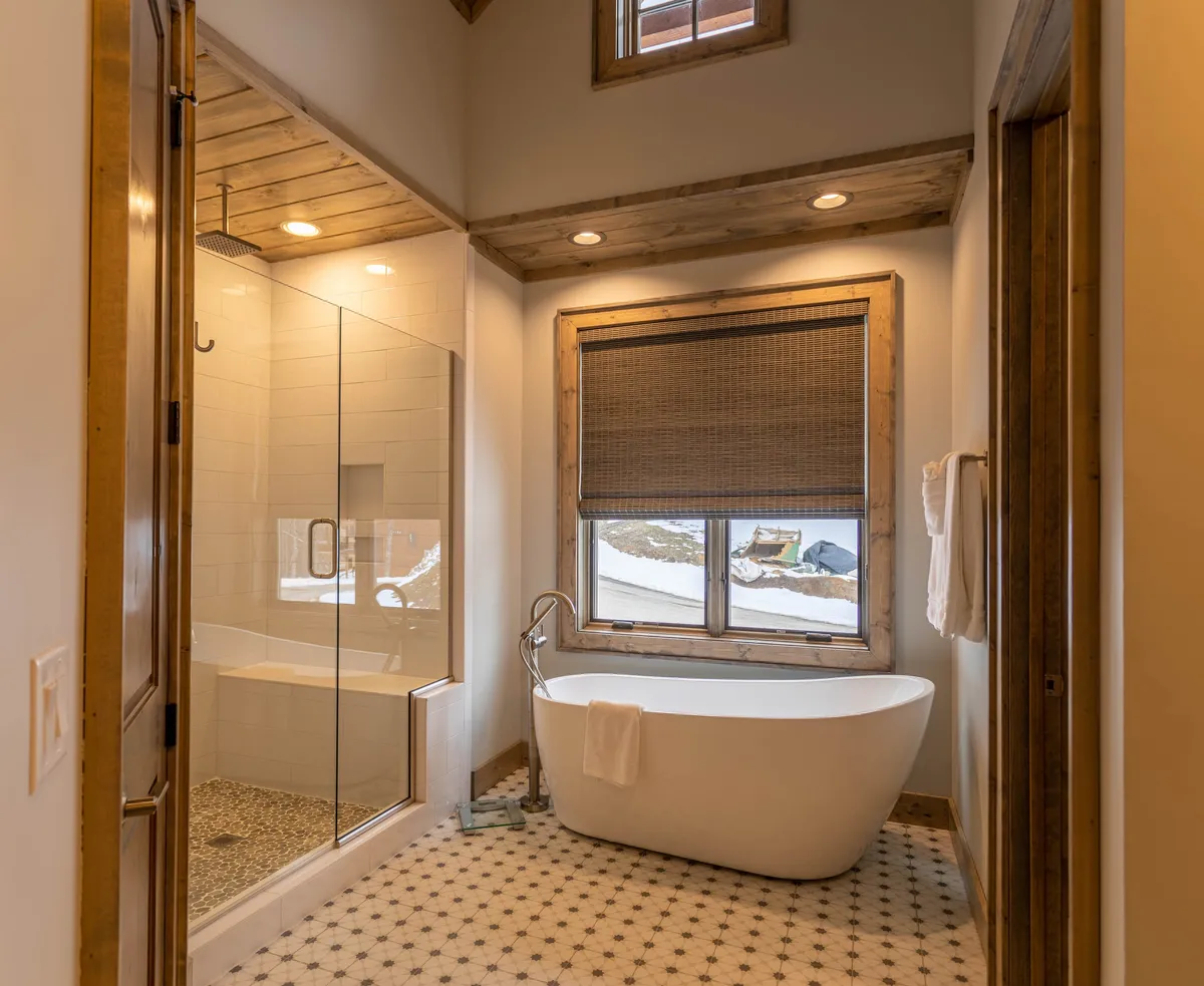 A luxurious bathroom featuring a freestanding soaking tub, a glass shower enclosure, wooden ceiling, and a window with a view. The space is filled with natural light and feels both spa-like and inviting.