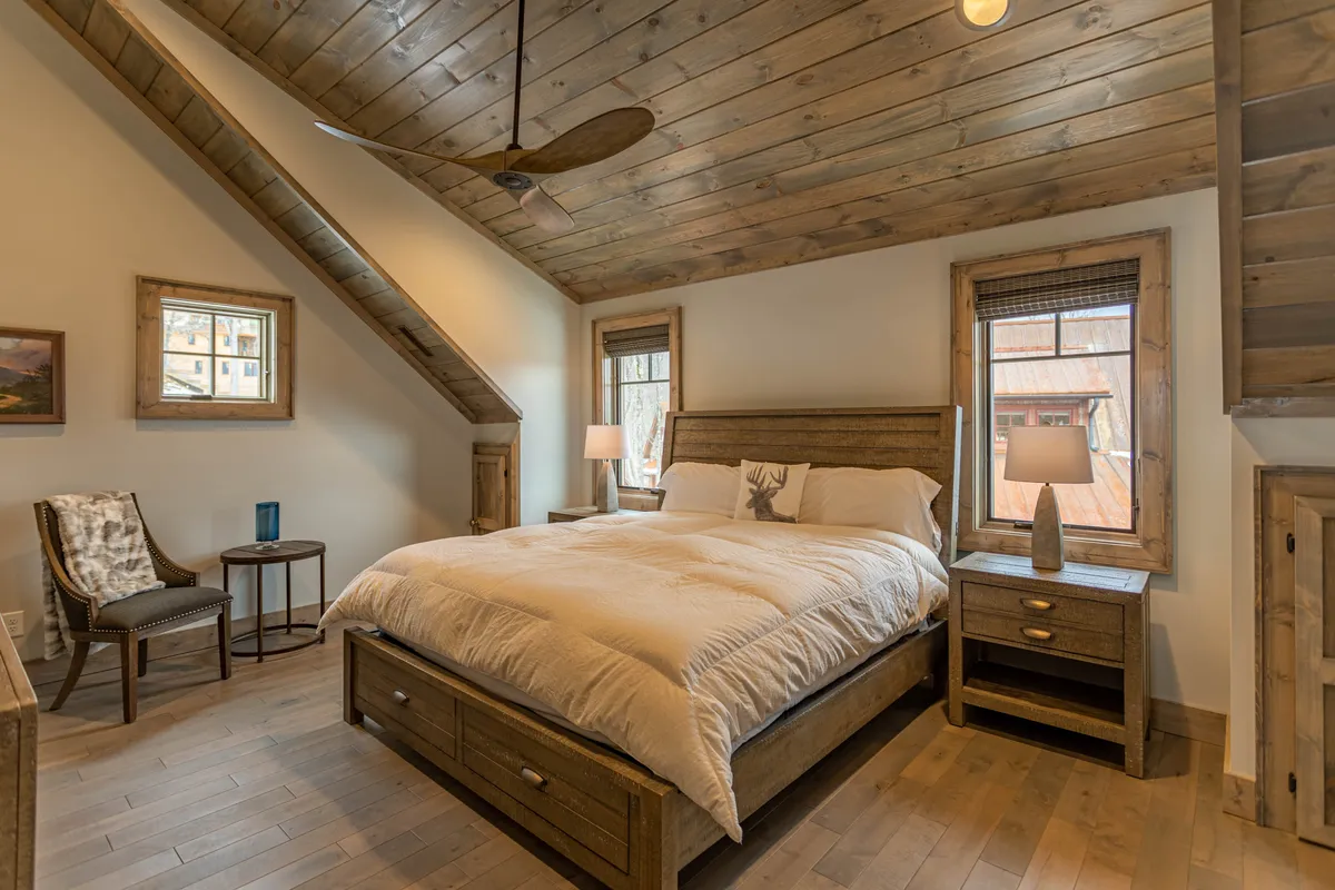 A serene king-sized bedroom with a wooden ceiling, large windows letting in natural light, nightstands, and a comfortable chair. The room exudes tranquility, blending plush bedding and soft lighting for a restful ambiance.