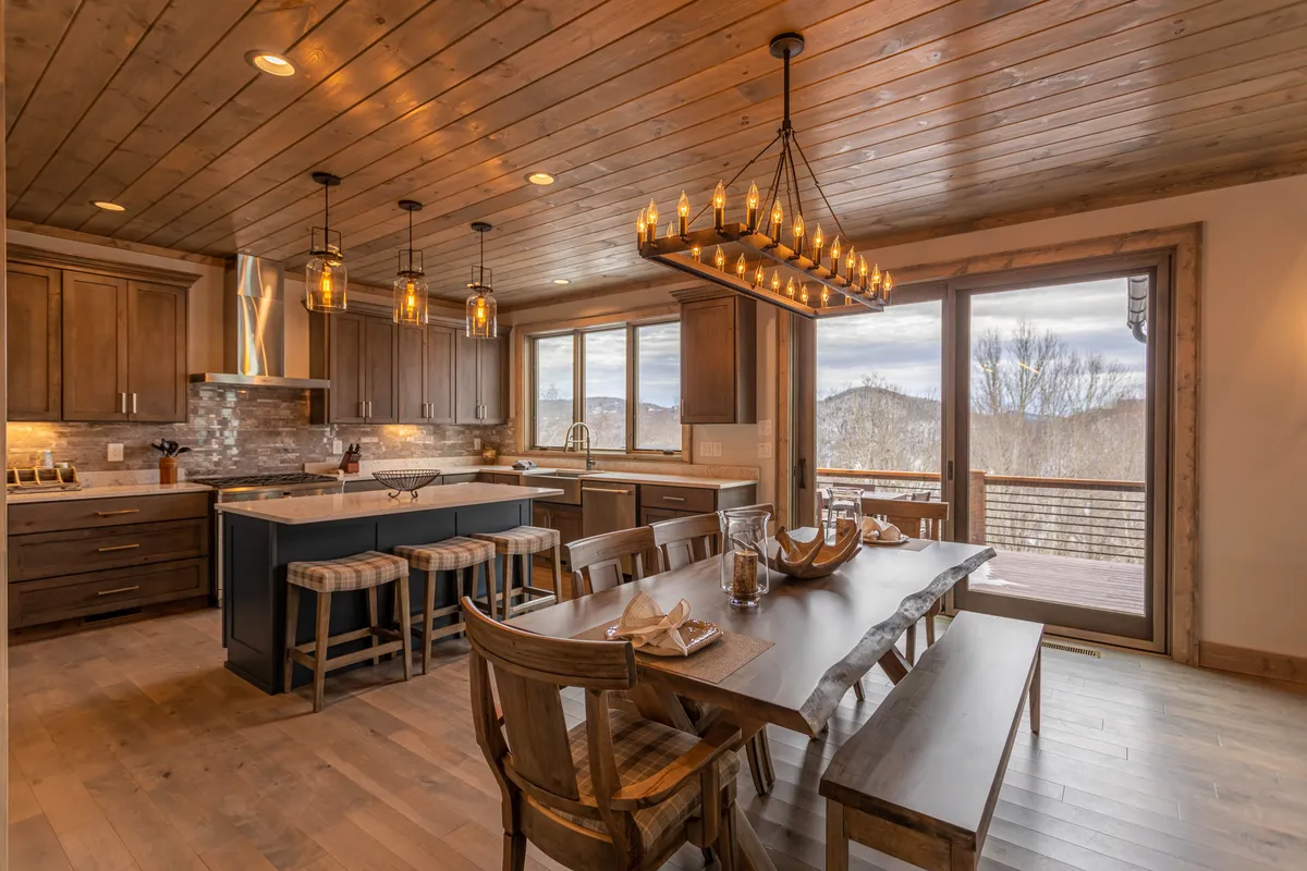 This image captures a spacious open-plan area featuring a large wooden dining table set for eight, a modern kitchen island, wooden cabinetry, a stylish chandelier, and panoramic windows that flood the space with natural light and offer scenic views. The kitchen is equipped with high-end fixtures, and the overall ambiance is one of warmth, comfort, and sophisticated functionality.