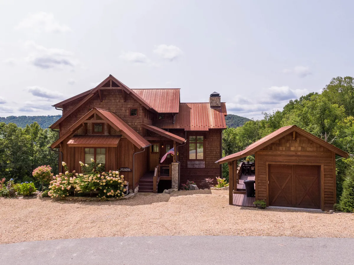 The image showcases the inviting wooden cabin with a striking red metal roof, surrounded by lush, flowering bushes. The spacious porch and garage hint at comfort, privacy, and mountain charm, all set against the tranquil backdrop of Banner Elk.