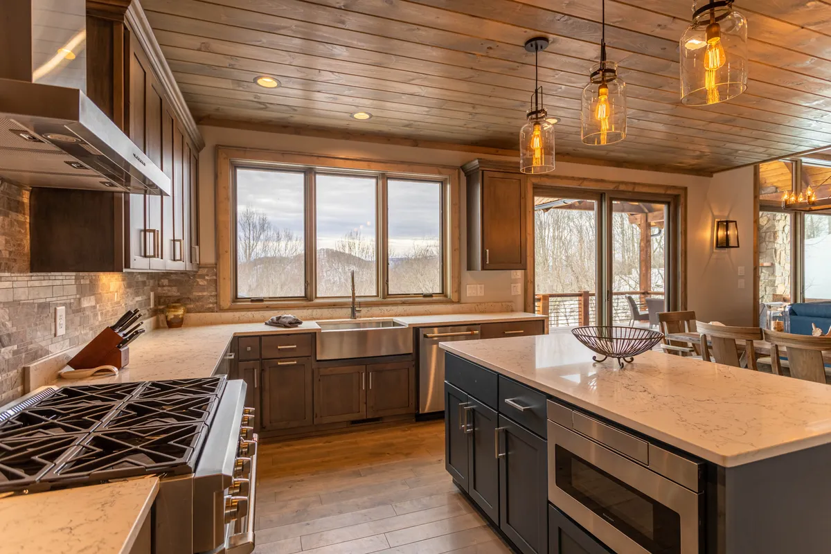 A modern kitchen and dining space with large windows framing stunning mountain views. The kitchen features a granite island, modern appliances, and stylish light fixtures. The open layout emphasizes spaciousness and effortless flow between cooking, dining, and enjoying the views outside.