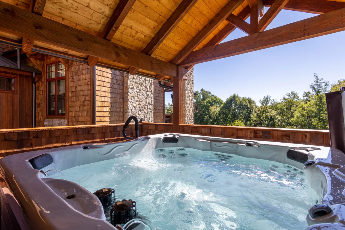 A tranquil outdoor hot tub surrounded by wooden beams, stonework, and green trees creates a relaxing setting. The area offers both privacy and a direct connection to nature, perfect for unwinding after a day of family adventures.